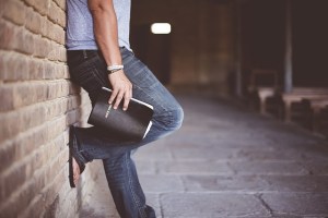 Lower body of person leaning on a brick wall with 1 foot on the wall and a Bible in hand