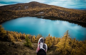 A person's tennis shoes are seen above a lake surrounded by trees and hills